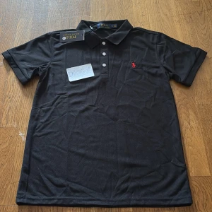 Svart pikétröja från Ralph Lauren - Klassisk svart pikétröja från Ralph Lauren med tre vita knappar och röd broderad logga på bröstet. Tröjan har korta ärmar och är tillverkad i mjuk bomull som känns skön mot huden. Perfekt för en clean och stilren look.