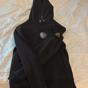Svart hoodie från Stone Island - Svart hoodie från Stone Island med klassisk logga på ärmen och bröstet. Tröjan har huva med snörning och känguruficka framtill. Tillverkad i 100% bomull för en skön och avslappnad känsla. Perfekt för dig som gillar streetwear och snygga detaljer.
