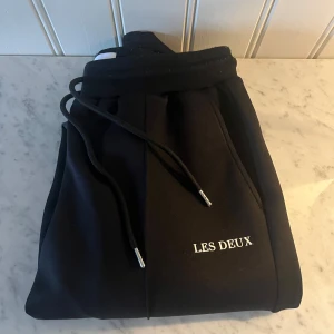 Svarta mjukisbyxor från Les Deux - Svarta mjukisbyxor från Les Deux med broderad vit logga på benet. Byxorna har snörning i midjan och är tillverkade i mjukt material som känns riktigt skönt. Passformen är normal och de har raka ben med diskreta sömdetaljer framtill. Köpt helt ny, för 1200! Kvitto finns.