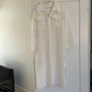 Vit lång skjortklänning i linne - Vit lång skjortklänning i luftig linneblandning med stora bröstfickor, klassisk krage och långa ärmar. Rak passform med slitsar i sidorna och stilren look. Perfekt för en chill och avslappnad vibe.
