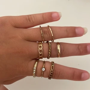 Set med åtta guldplaterade ringar - Säljer ett set med åtta trendiga guldiga ringar i olika former och stilar. Några har kedjelänk-design, en är tvinnad och en har en liten glittrande sten. Perfekt för att mixa och matcha på flera fingrar för en cool look.