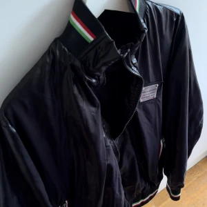 Svart bomberjacka från Dolce & Gabbana - Svart bomberjacka från Dolce & Gabbana med glansig finish och ribbade muddar i svart med röd, vit och grön rand. Storlek S men passar även Xs, blivit för liten för mig så därför säljer jag! Ca 2 år gammal! Ser ut som Italiensk Mafia i denna!