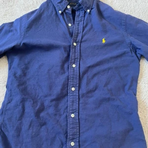 Blå skjorta från Polo Ralph Lauren - Klassisk blå skjorta från Polo Ralph Lauren i slim fit-modell. Skjortan har långa ärmar, knappar framtill och ikoniska gula loggan broderad på bröstet. Tillverkad i mjuk bomull och passar perfekt till jeans eller chinos. Varan skickas dagen vid köp!💸