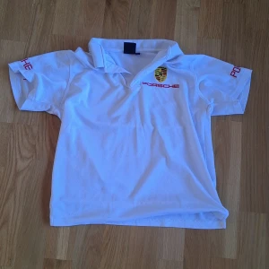 Vit Porsche piké med broderad logga - Vit piké från Porsche med klassisk krage och broderad Porsche-logga på bröstet och ryggen. Röd text på ärmarna och framtill. Skön passform och sportig look, perfekt för dig som gillar bilar och snygga märkeskläder. Pris kan såklart diskuteras!