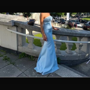 Ljusblå axelbandslös balklänning - Säljer en elegant ljusblå långklänning med axelbandslös design och figurnära passform. Klänningen har glittriga detaljer upptill och en lätt utsvängd nederdel som ger en snygg siluett. Perfekt för bal eller student!