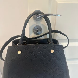 Svart handväska i skinn - Säljer en svart handväska i präglat skinn. Väskan har både handtag och axelrem, guldfärgade metalldetaljer, dragkedja och ett hänglås. Perfekt för dig som vill ha en lyxig och rymlig väska med ikonisk design. Funkar även till skolan