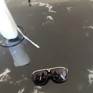 Svarta Louis Vuitton solglasögon perfekt till sommaren  - Snygga svarta solglasögon från Louis Vuitton med guldiga detaljer och klassisk pilotform. Glasögonen har mörka linser och stilrena skalmar med Louis Vuitton-logga i guld. Perfekt accessoar för att lyfta din look.