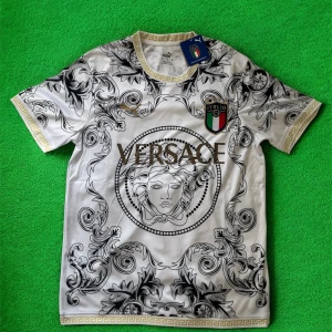 Italien x Versace x Puma fotbollströja - Snygg vit fotbollströja i samarbete mellan Italien, Versace och Puma. Tröjan har korta ärmar, guldiga detaljer och ett unikt svart barockmönster över hela plagget. Materialet är glansigt och lätt, perfekt för dig som vill sticka ut på planen.