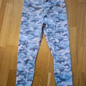  camoflerade  leggings från Fabletics  - Snygga leggings med rosa och grått camouflagemönster. Hög midja med korsad band baktill ger en cool detalj. Stretchigt material som sitter tajt och bekvämt, perfekt för träning eller chill.