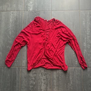 Röd omlottkofta med långa ärmar - Säljer en röd omlottkofta med långa ärmar. Koftan är tunn och har en avslappnad passform, perfekt att slänga över en topp. Vet ej storlek men runt S/M