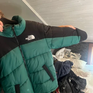 Grön pufferjacka från The North Face - Säljer en grön och svart pufferjacka från The North Face med hög krage och dragkedja framtill. Jackan har två snedställda fickor med svarta detaljer och broderad logga på bröstet. Perfekt för kalla dagar och riktigt snygg streetstil.