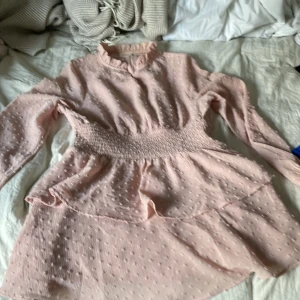 Ljusrosa kortklänning från Gina Tricot - Supersöt ljusrosa kortklänning från Gina Tricot med långärmade ärmar, smock i midjan och volangdetaljer. Klänningen har diskret prickigt mönster i tyget och en liten volangkrage som ger en romantisk vibe. Perfekt för dig som gillar en feminin och trendig stil.