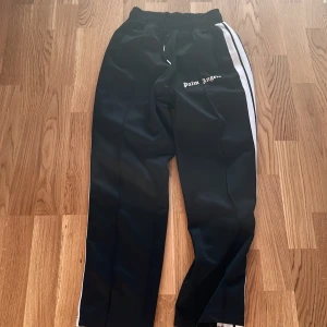 Palm Angels svarta trackpants med vit stripe | 170/S - Svarta trackpants från Palm Angels med vit sidoremsa och logga tryckt på ena benet. Byxorna har elastisk midja med snörning och är tillverkade i ett glansigt syntetmaterial. Perfekta för en sportig och trendig streetwear-look.