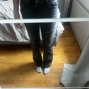 Bootcut svarta jeans med slitning - Säljer ett par svarta bootcut jeans med slitning över knät. Jeansen har låg midja och klassisk femficksmodell. Materialet är denim och passformen är normal med lätt utsvängda ben. Perfekta för dig som gillar en avslappnad och trendig look.