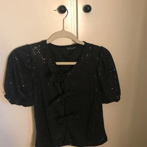 Glittrig svart topp från Missay - Svart glittrig topp från Missay med kort puffärm och tre söta rosetter framtill. Toppen har v-ringning och är figurnära utan att vara tight. Perfekt för dig som vill sticka ut med lite extra glitter och coola detaljer.