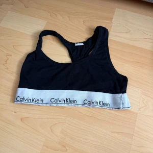 Svart topp från Calvin Klein - Svart topp från Calvin Klein med bred, grå resår nertill där loggan är tryckt flera gånger. Modellen är ärmlös med brottarrygg och passar perfekt till träning eller chill. Mjuk bomullskänsla och sportig look.