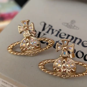 Vivienne Westwood guldörhängen med kristaller - Ikoniska Mayfair Bas Relief örhängen från Vivienne Westwood i guldtonad mässing med glittrande AB-kristaller. De har den klassiska orb-formen med detaljerade nitar och färgskiftande stenar som fångar ljuset. Perfekta för dig som vill sticka ut med statement-smycken.