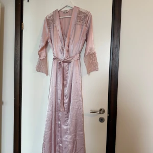 Ljusrosa långklänning i satin med spets - Superfin ljusrosa långklänning i satin med tunna axelband och v-ringning. Klänningen har vackra spetsdetaljer framtill och baktill samt en liten rosett vid bysten. Tillhörande matchande kimono med spets på ärmarna och knytband i midjan.