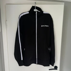 Palm Angels tracksuit, Svart L - Svart tracksuit från Palm Angels med vita ränder längs sidorna och logga tryckt på låret. Byxorna har elastisk midja och dragkedjor vid bensluten för extra stilpoäng. Perfekta för en chill och sportig look.
