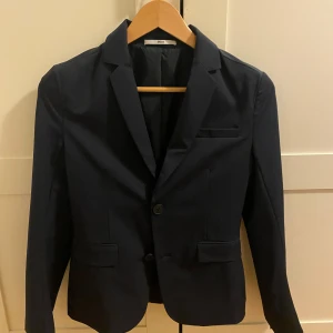 Mörkblå kavaj från Hugo Boss  - Snygg mörkblå kavaj från Hugo Boss i slim fit-modell. Klassisk design med två knappar framtill, tre fickor och diskreta knappar vid ärmslutet. Perfekt för dig som vill ha en stilren och modern look. Fodrad insida med små prickar för extra detalj.