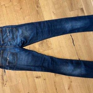 Blåa jeans från Nudie Jeans - Säljer ett par mörkblå jeans från Nudie Jeans med klassiska kontrastsömmar och snygga detaljer på bakfickorna. Jeansen har en smal passform och är tillverkade i slitstarkt denim. Perfekta för dig som gillar en clean och stilren look. Haft på dom en gång och tvättats två gånger.