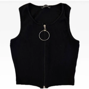 Svart ribbad linnetopp med dragkedja - Svart ribbad linnetopp från New Favori Collecion med en cool silverfärgad dragkedja framtill och stor ring som detalj. Ärmlös och croppad modell som ger en edgy vibe. Perfekt att styla med höga jeans eller kjol.