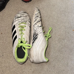 Adidas vita fotbollsskor med limegrönt - Snygga vita Adidas fotbollsskor med svarta ränder och limegröna snören samt innersula. Skorna har svart mönster på ovandelen och är tillverkade i syntetmaterial. Perfekta för dig som vill sticka ut på planen!