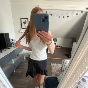 Svart volangkjol från UNIQ YOUNG - Supersöt svart kortkjol från UNIQ YOUNG med volangdetaljer och lätt glansigt tyg. Kjolen har elastisk midja och en omlottliknande design framtill. Perfekt för dig som vill ha en trendig och bekväm kjol med lite extra flärd.