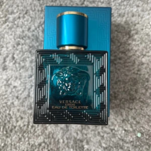 Versace Eros Eau de Toilette - Versace Eros Eau de Toilette i en lyxig turkosblå glasflaska med guldiga detaljer och ikonisk Medusa-logga framtill. Flaskan har en stilren, fyrkantig form och ett matchande blått lock med guldkant. En klassisk och kraftfull doft för dig som gillar statement-parfymer.