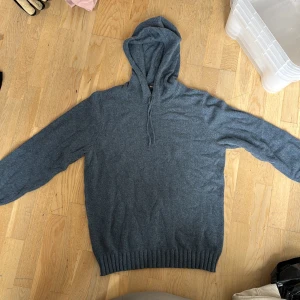 Grå stickad hoodie från Dressmann - Mysig grå stickad hoodie från Dressmann i storlek L. Tröjan har huva med snörning och ribbade muddar vid ärmslut och nederkant. Perfekt för dig som gillar en chill och stilren look under höst och vinter.