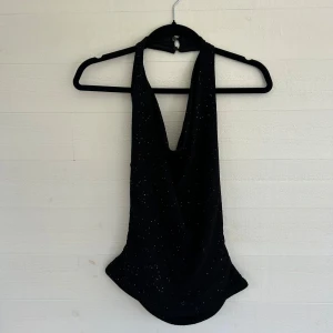 Svart glittrig halterneck topp NA-KD - Supersnygg svart halterneck topp från NA-KD med glittriga detaljer. Toppen har en draperad framsida och är ärmlös med djup urringning. Perfekt för dig som vill sticka ut med lite extra glitter. Materialet känns mjukt och stretchigt.