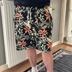 Blommiga svarta shorts med snörning - Snygga svarta shorts med färgglatt blommönster i grönt, vitt och orange. De har en bekväm midja med vit snörning och avslappnad passform, perfekta för varma dagar. Shortsen har sidofickor och är tillverkade i ett lätt material som känns skönt mot huden.