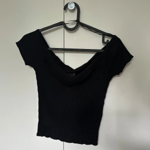 Svart ribbad topp Gina Tricot - Svart ribbad topp från Gina Tricot med bred v-ringning som går offshoulder. Toppens material är stretchigt och sitter tight mot kroppen. Perfekt för dig som gillar en enkel men snygg stil. Oanvänd med lappen kvar!
