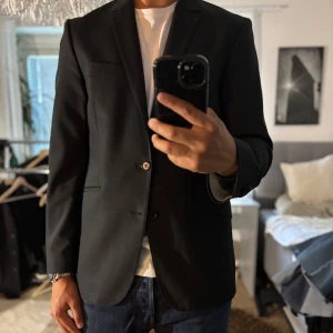 Svart kavaj från SIR of Sweden - Stilren svart kavaj från SIR of Sweden i klassisk modell med två knappar och smala slag. Tillverkad i en mix av polyester och ull med lite lycra för extra komfort. Perfekt för dig som vill ha en snygg och tidlös look.