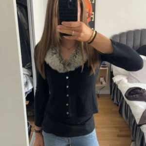 Svart kofta med fuskpäls från KappAhl - Svart kofta från KappAhl med bred, grå fuskpäls-krage och knappar framtill. Koftan har långärmad passform och är tillverkad i en mix av ull och syntet. Perfekt för dig som vill ha en mysig och snygg look med lite extra edge. Obs storleken stämmer ej. Passar S-M