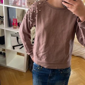Lila sweatshirt med pärldetaljer - Superfin brun sweatshirt med långa ärmar och coola pärldetaljer på axlarna. Tröjan har en relaxed passform och är gjord i mjuk bomull, perfekt för dig som vill ha något extra till din outfit. Rund halsringning och ribbade muddar.