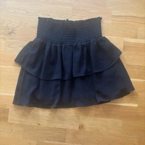 Svart volangkjol från Made in Chelsea - Supersöt svart kortkjol från Made in Chelsea med smockad midja och dubbla volanger.