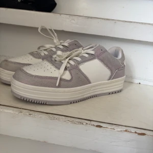 Vita och ljuslila sneakers med snörning - Snygga sneakers i vitt och ljuslila med klassisk låg profil. Skorna har snörning, perforerade detaljer på tån och en platt sula. Materialmix av textil och syntet ger en fräsch och modern look, perfekt för dig som gillar stilrena och trendiga skor.