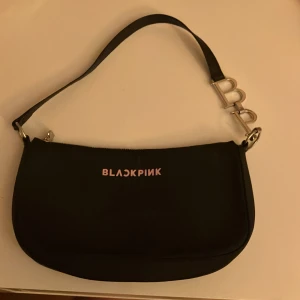 Svart axelväska BLACKPINK med BP-detalj - Snygg svart axelväska från BLACKPINK med rosa logga framtill. Väskan har en silvrig dragkedja och coola metallbokstäver 'B' och 'P' på axelremmen. Tillverkad i slätt syntetmaterial med svart foder. Perfekt för dig som vill sticka ut med K-pop vibes.