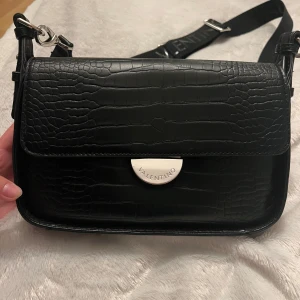 Valentino väska  - Snygg svart axelväska från Valentino med croco-mönstrad yta och silverfärgade detaljer. Väskan har justerbar axelrem med logga, praktiska fack med dragkedja både bak och inuti samt stilren metallknäppning framtill. Har några få defekter på några av de silverfärgade delarna. Kan ej lägga upp fler bilder. Vid intresse skickar jag tydligare bilder på defekterna. 