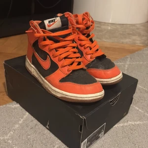 Nike Dunk High svart och orange - Nike Dunk High sneakers i svart och orange med klassisk siluett. Skorna har snörning, rund tå och vit sula. Ovandelen är i syntet och textil, med orange detaljer och swoosh. Perfekta för dig som gillar streetwear och vill sticka ut.