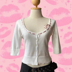 Japansk vit kofta med rosett från ofuon palette - Supersöt japansk vit kofta från ofuon palette med trekvartsärm och knappar framtill. Urringningen är dekorerad med ett rosa satinband och en liten rosett på ena sidan. Modellen är croppad och har ribbade muddar för en extra snygg passform. Importerad från Japan. 