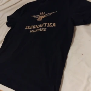 Svart Aeronautica Militare t-shirt - Svart t-shirt från Aeronautica Militare med snyggt tryck av en örn och text på bröstet. Klassisk passform och rund halsringning, perfekt för dig som gillar stilrena och coola plagg med militärinspirerad vibe.