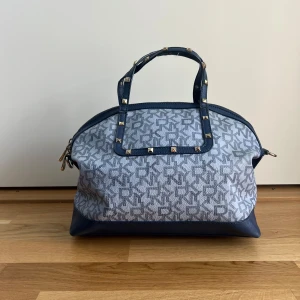 Blå axelväska från DKNY med nitar - Snygg axelväska från DKNY i blått och grått med logomönster över hela väskan. Väskan har både handtag och justerbar axelrem i blått skinn, samt coola guldfärgade nitar på handtaget. Stängs med dragkedja och har detaljer i guldmetall. Ungefärliga mått (platta): Längs botten: 31 cm Topp till botten exklusive handtag: 25 cm Topp till botten inklusive handtag: 35 cm Sida till sida längst ner: 15 cm Sida till sida i mitten: 25 cm
