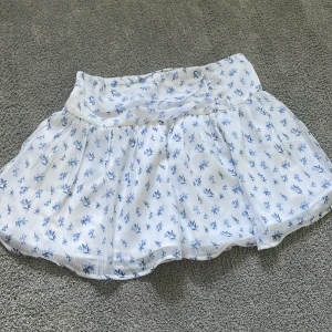 Vit blommig kortkjol från Gina Tricot - Supersöt vit kortkjol från Gina Tricot med blått blommönster. Kjolen har en lätt utsvängd form och bred midja för en bekväm passform. Perfekt för varma dagar och ger en fräsch sommarkänsla.