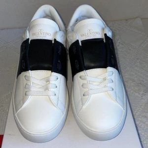 Valentino open skor - Snygga Valentino Garavani Open sneakers i vitt skinn med bred svart läderdetalj över vristen. Klassisk rund tå, vita snören och diskreta nitar på hälen. Perfekta för dig som vill ha en stilren och lyxig look. Priset kan diskuteras vid snabb affär 