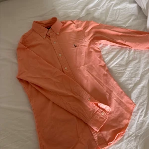 Korallfärgad skjorta från Ralph Lauren - Snygg korallfärgad skjorta från Ralph Lauren i slim fit-modell. Skjortan har klassisk krage, knäppning framtill och broderad logga på bröstet. Tillverkad i bomull och har långa ärmar med knappar vid manschetterna. Perfekt för dig som gillar stilrena plagg med en twist.
