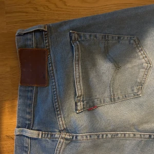 Levi's 501 ljusblå jeans straight fit - Klassiska Levi's 501 jeans i ljusblå tvätt med raka ben och femficksmodell. Jeansen har den ikoniska läderpatchen bak och röd Levi's-tagg på bakfickan. Tillverkade i slitstarkt bomullsjeans, perfekt för en avslappnad och tidlös stil.