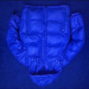 Blå dunjacka från Polo Ralph Lauren - Säljer en blå dunjacka från Polo Ralph Lauren med huva och dragkedja. Jackan har två sidofickor med dragkedja och den klassiska röda Polo-loggan på bröstet. Perfekt för kalla dagar och riktigt snygg till streetwear-stilen.