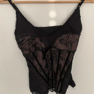korsett-topp - Supersnygg svart korsett-topp med tunna axelband, transparent mesh och broderade blommor med glittriga detaljer. Toppen har spetskant, liten rosett framtill och figurnära passform. 